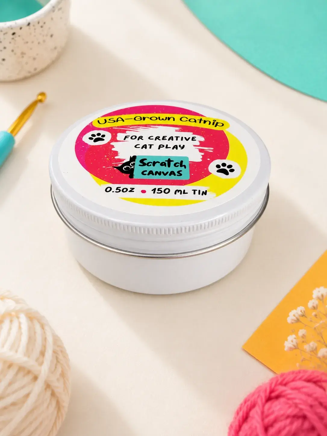 Closed Scratch Canvas Catnip Tin on cream paper in an artist-studio setting, with balls of yarn and a crochet hook around it.