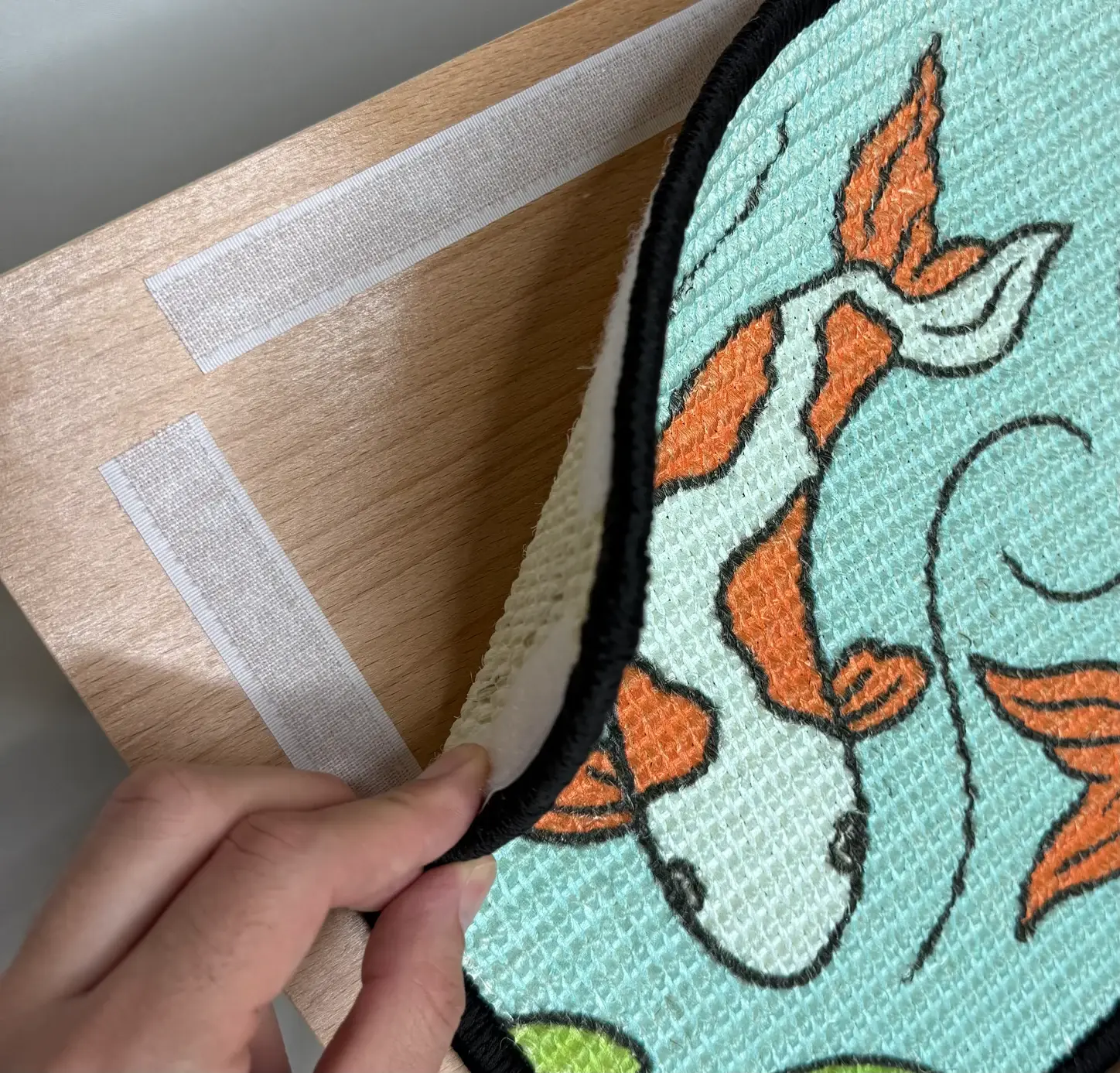 Hand lifting the Koi scratch panel from the wooden easel, revealing the hook-and-loop fastening strips underneath.